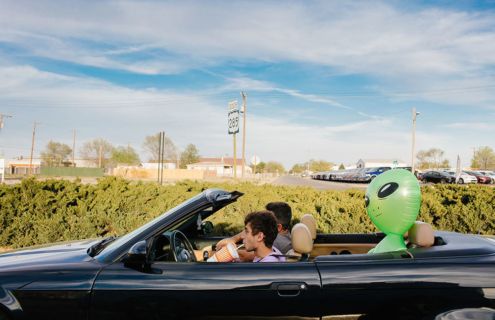 A picture of two people in a convertible car with a blow-up alien in the backseat