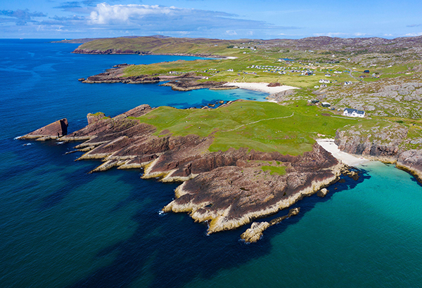 This aerial view shows the coastal cliffs at Clachtoll in northwest Scotland. The giant ancient boulder sits along the shore on the right side of the image, forming a hill along the small beach in front of the building.
