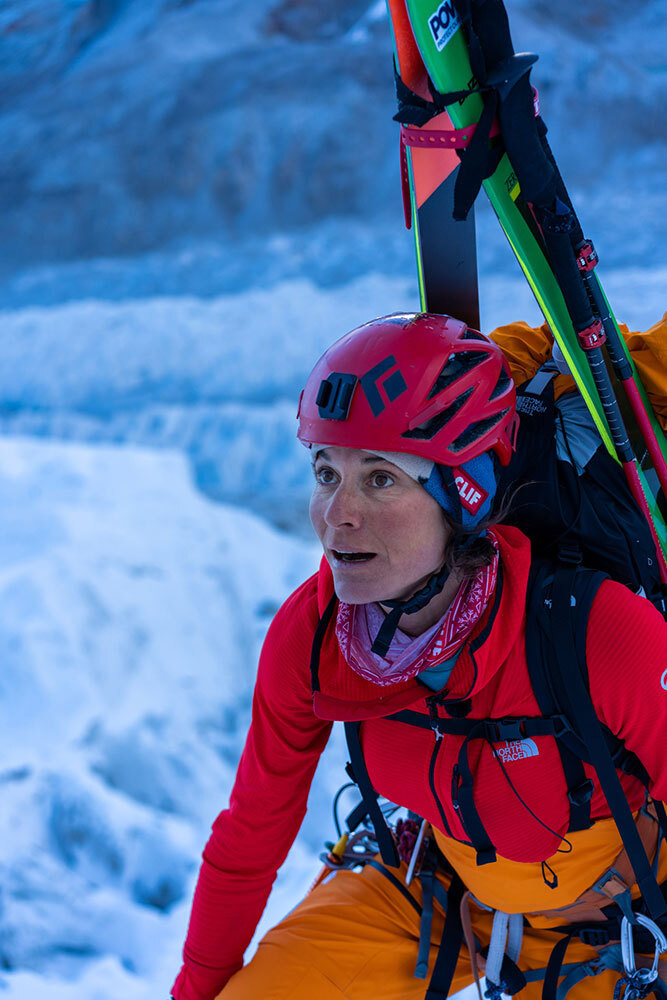 A woman wearing a helmet and carrying skis climbs an icy mountain