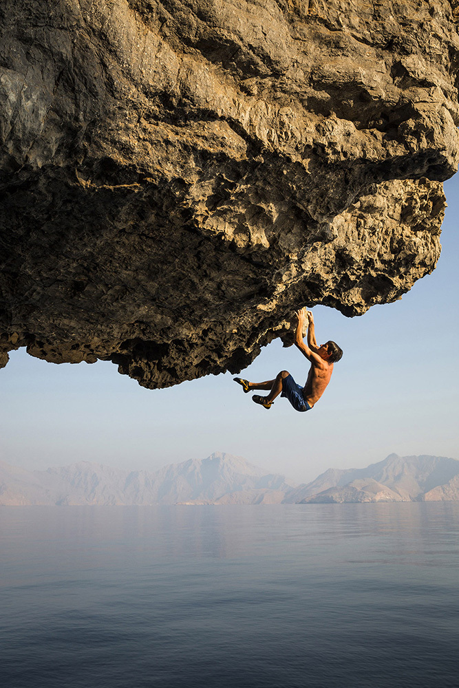 A sun-drenched climber, Alex Honnold, dangles from an overhang against a background of the ocean and distant land.