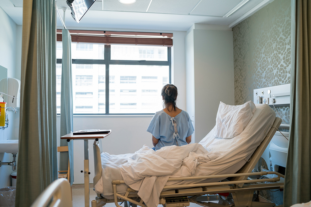 A woman wearing a hospital gown sitting up in a hospital bed and looking out a window.