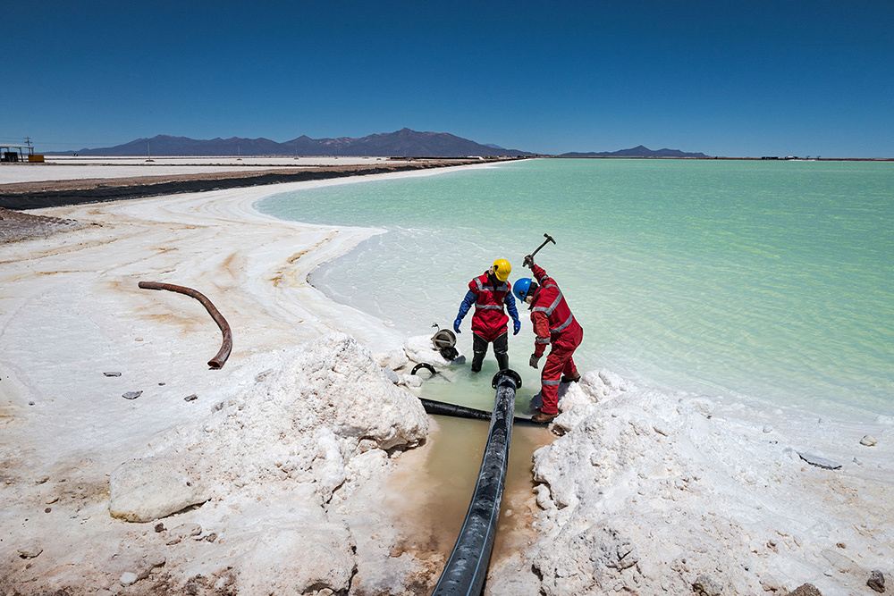 Workers, wearing red, one piece suits, at the lithium pilot plant use hammers to break up a layer of salt that periodically clogs the pipes transporting brine with lithium into large, turquoise blue evaporation pools.