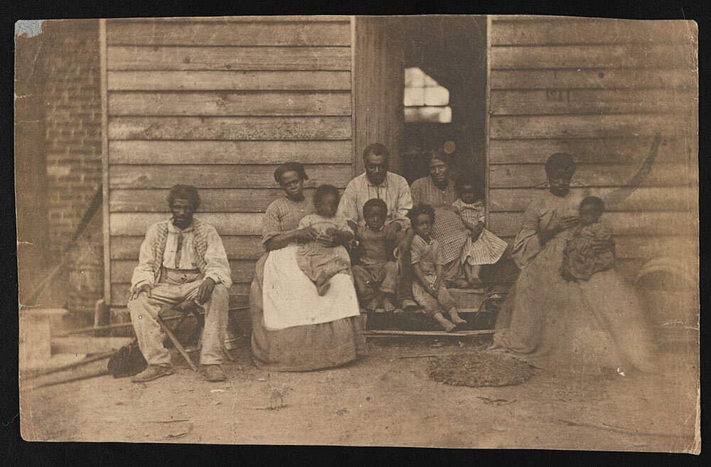 This historical photograph shows an enslaved African American family or families posing in front of a wooden house on a plantation in Hanover County, Virginia.