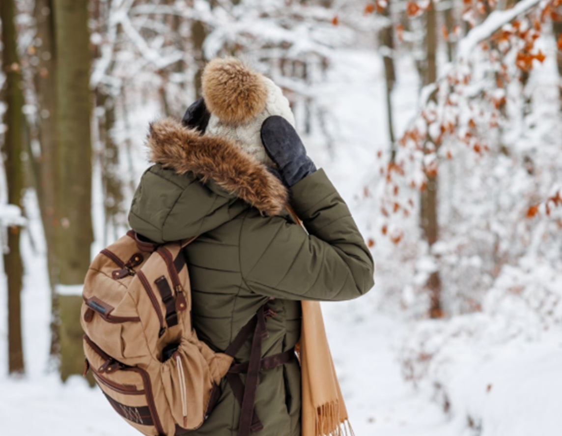 A woman wearing an olive green down jacket and tan backpack is seen from behind while standing in snow-covered woodlands. She is adjusting her white knitted hat with mitten covered hands.