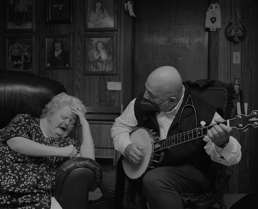 A doctor plays a banjo next to a patient in an armchair