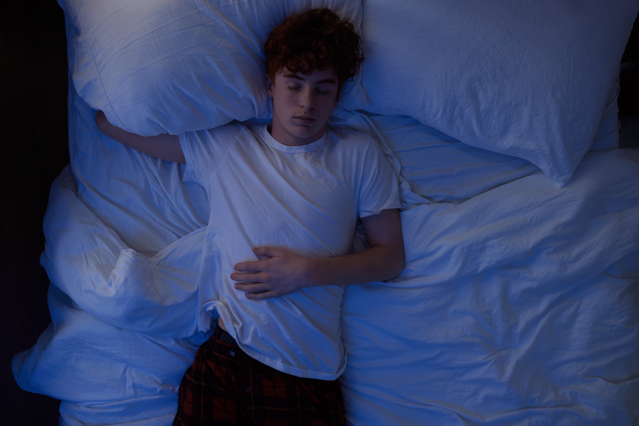 A teenage boy sleeping in bed, photographed from directly overhead while sleeping on his side, back, and stomach