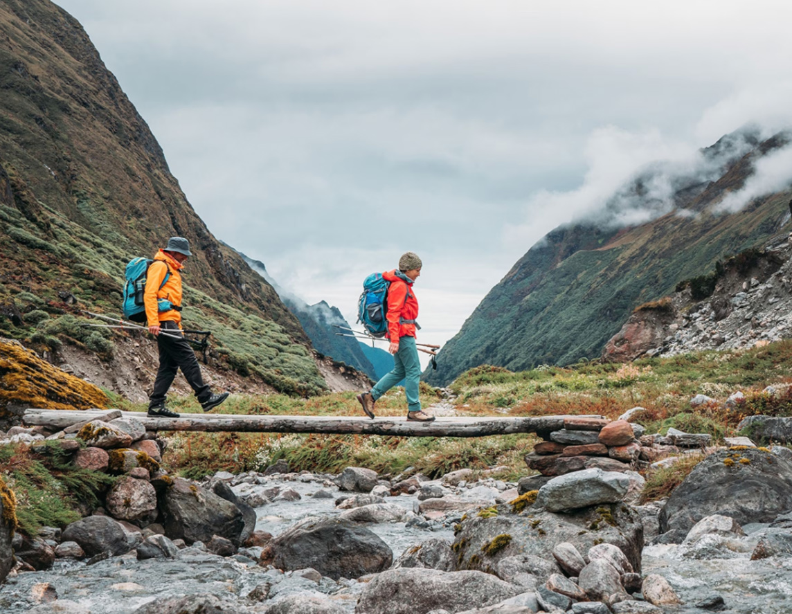 Two people walking across wood log over a stream of water.