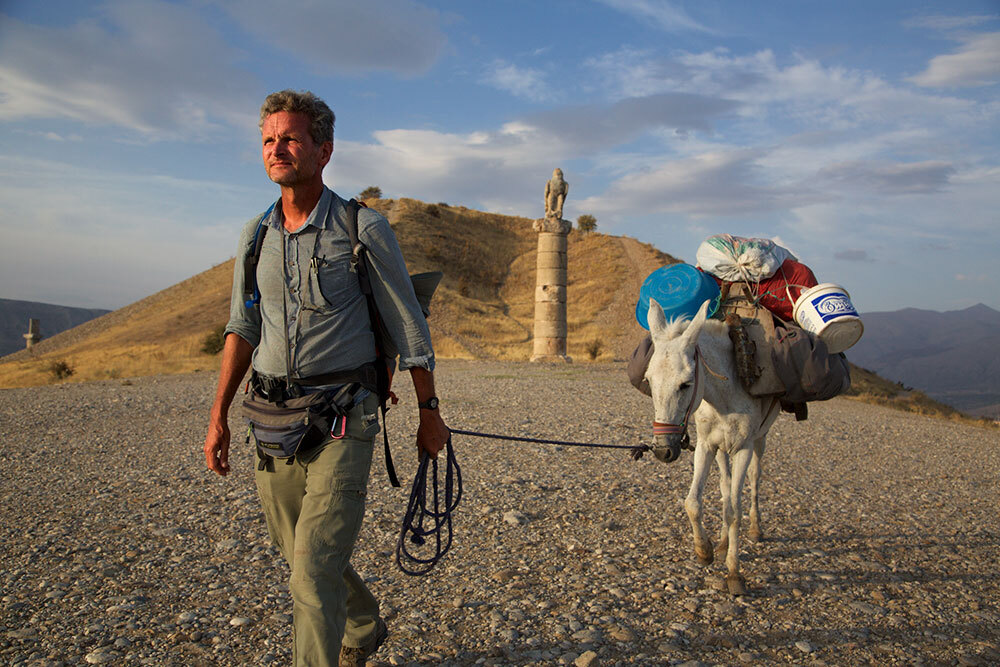 a man walking in an arid landscape in Turkey, puling a white mule carrying his belongings