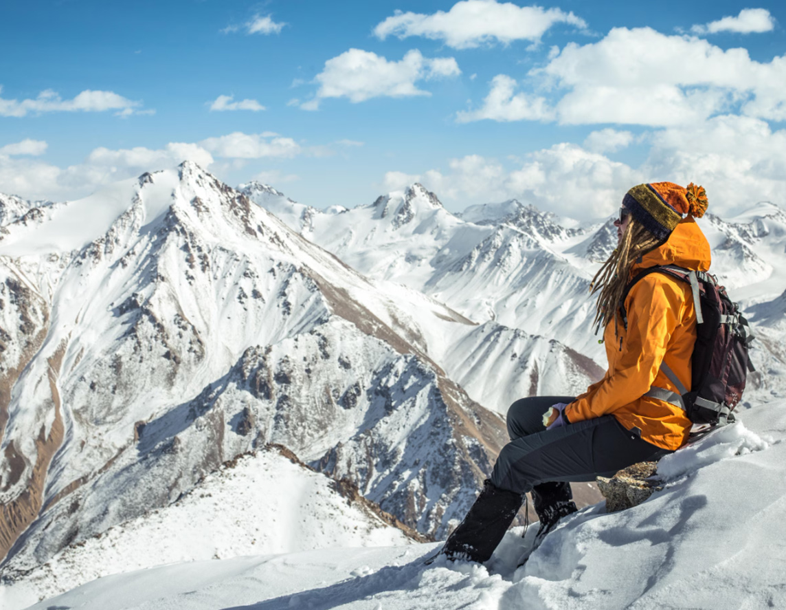 Outdoor girl is sitting on the top of mountain and enjoying the view on surroundings snow peaks.