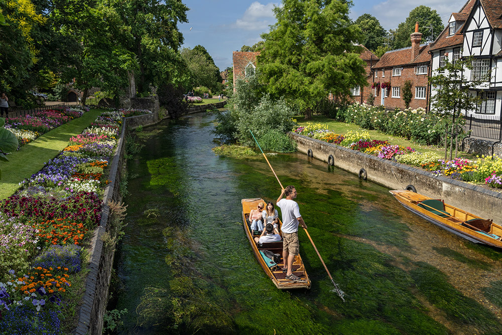 A person is standing on a little wooden boat with a large wooden pole while three people are seated enjoying the view