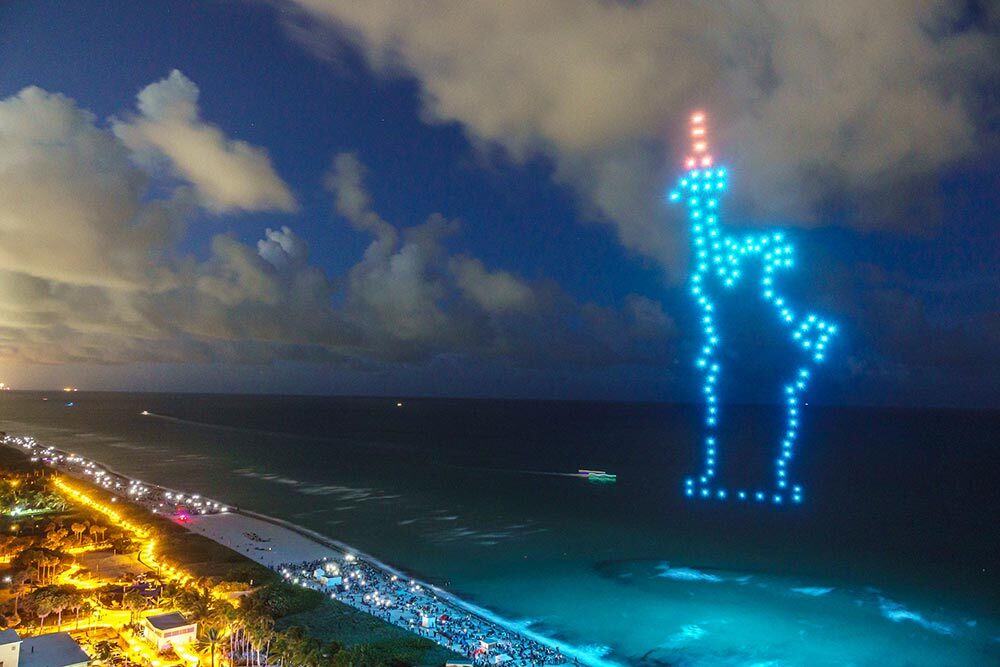 drone fireworks in the shape of the statue of liberty over the ocean near a beach.