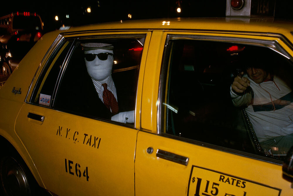 A man wrapped in bandages while wearing a suit sits in the back of a taxi.