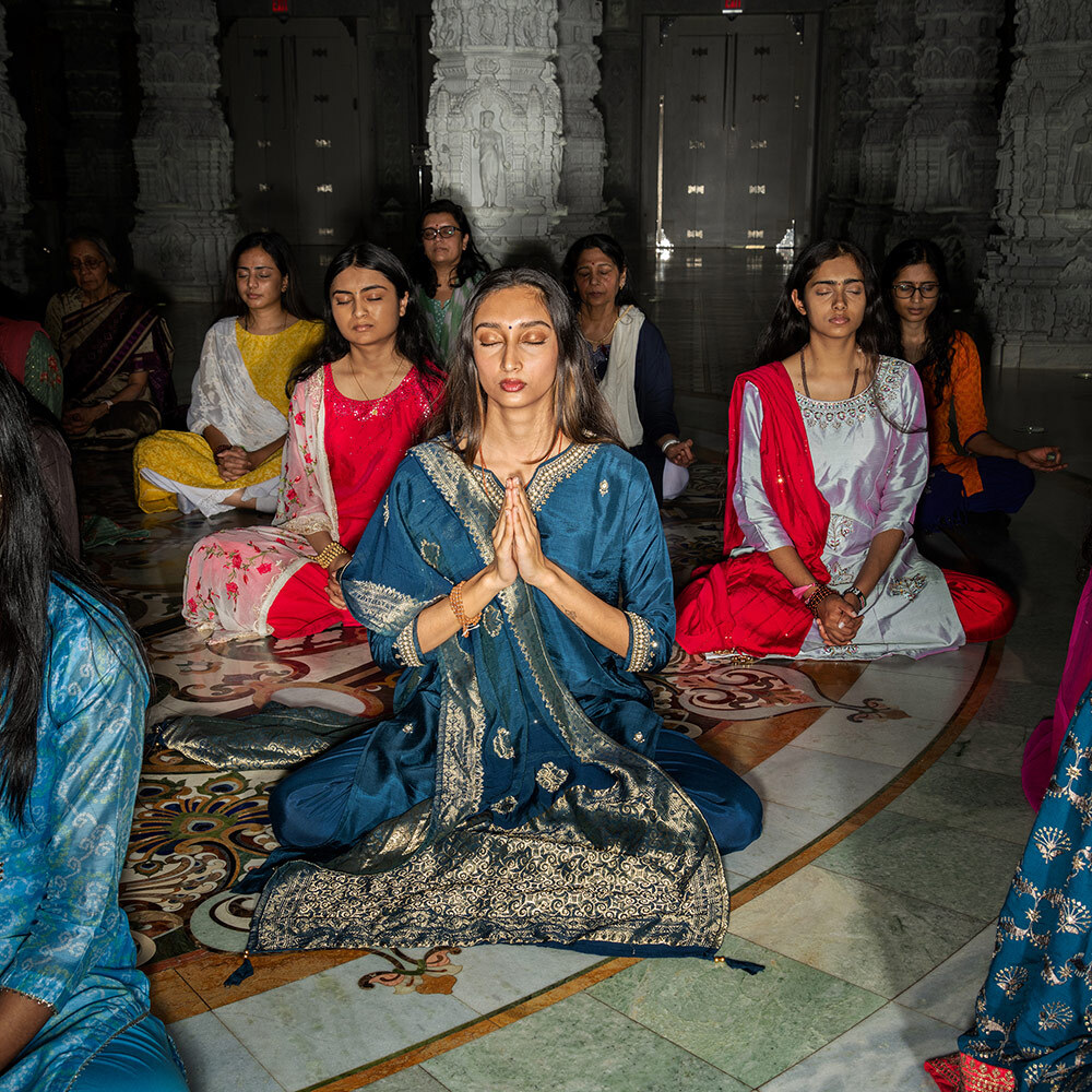 Numerous women wearing colorful garments sit cross-legged with their eyes closed inside of a large Hindu temple