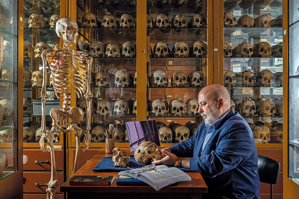 A man holds a skull at a desk. Shelves lined with skulls are in the background