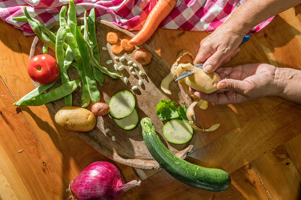 An overhead view of hands peeling a potato over a cutting board covered with various vegetables