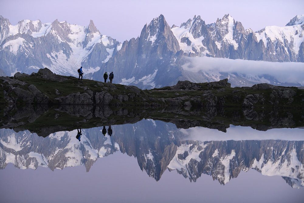 People walk along a lake with a mountain range in the background.