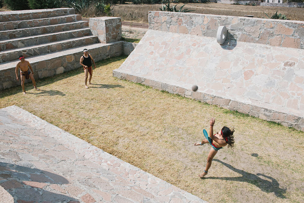 Three people play a game with a ball on a grassy court surrounded by stone walls