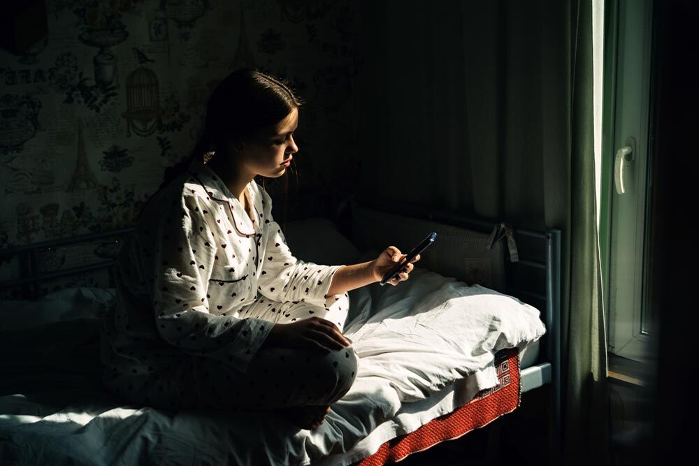 A teenage girl with mobile phone sitting on a bed in a mostly dark room. Bright, high contrast light shines in from a nearby window, illuminating part of the girl and her phone.