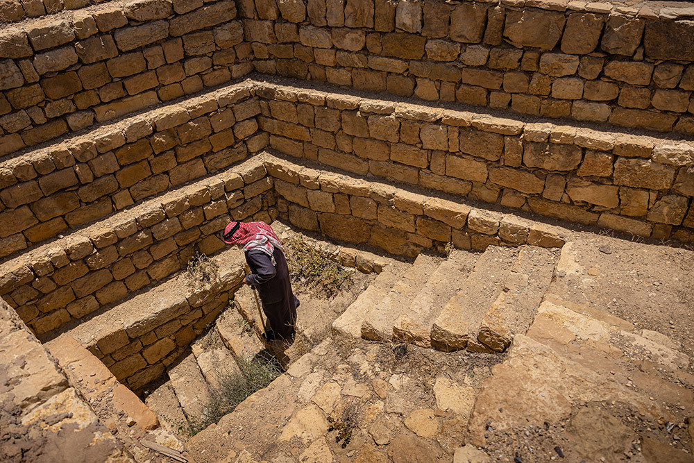 A man, with a red headdress, grey robe and cane, walks down stairs surrounded by layers of wall stones