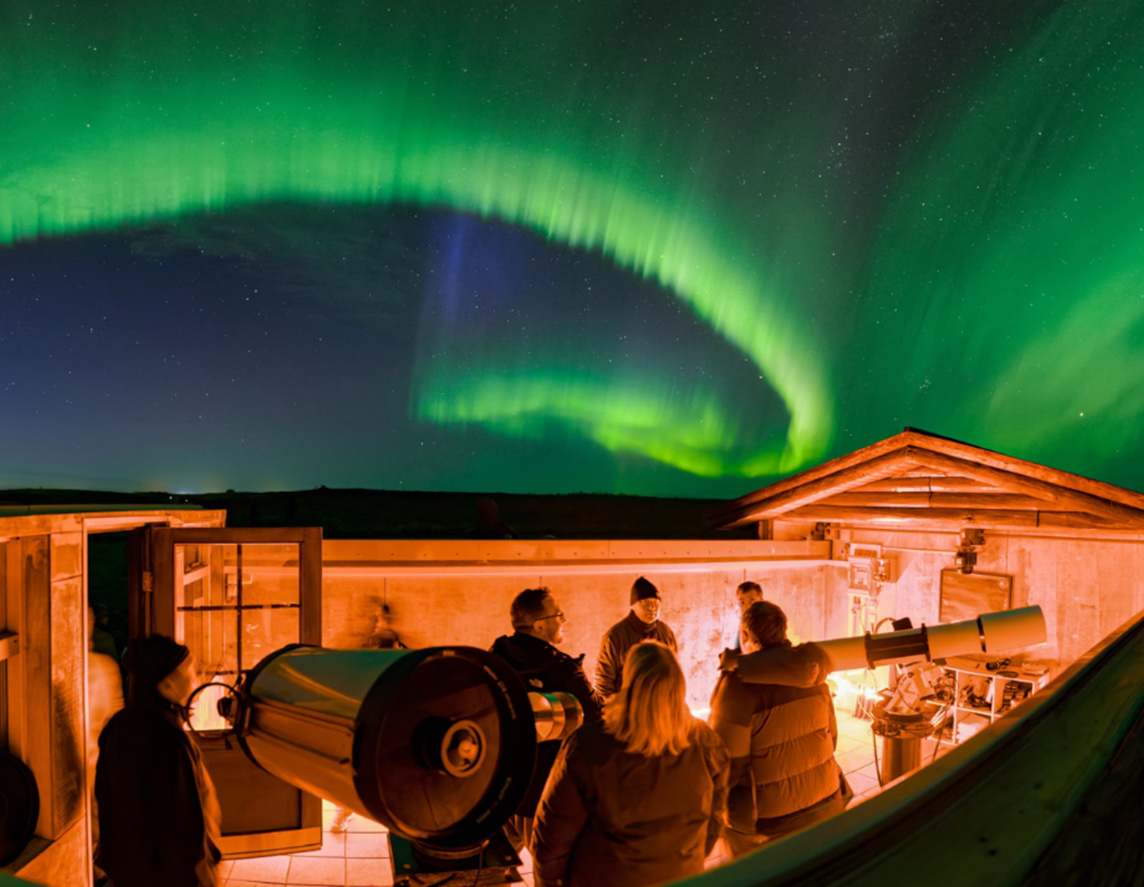 A group of people stand on a roof with telescopes, beneath green ribbons of the Northern Lights streaking across the sky.