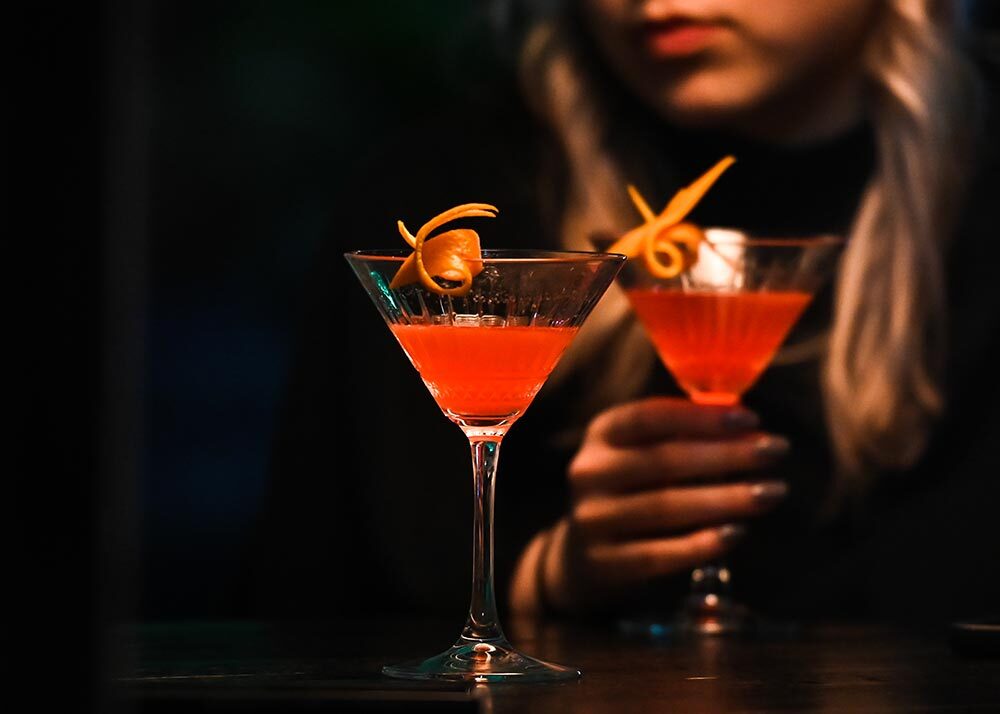 A cocktail on a table and a woman holding another cocktail in the background