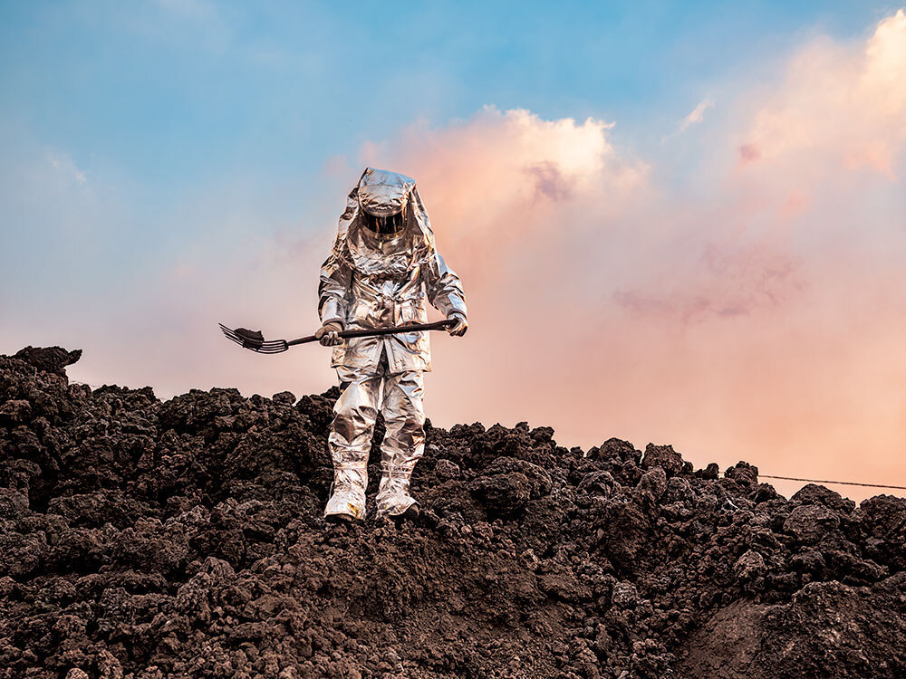 A person in a silver suit holds a pitchfork on lava rocks