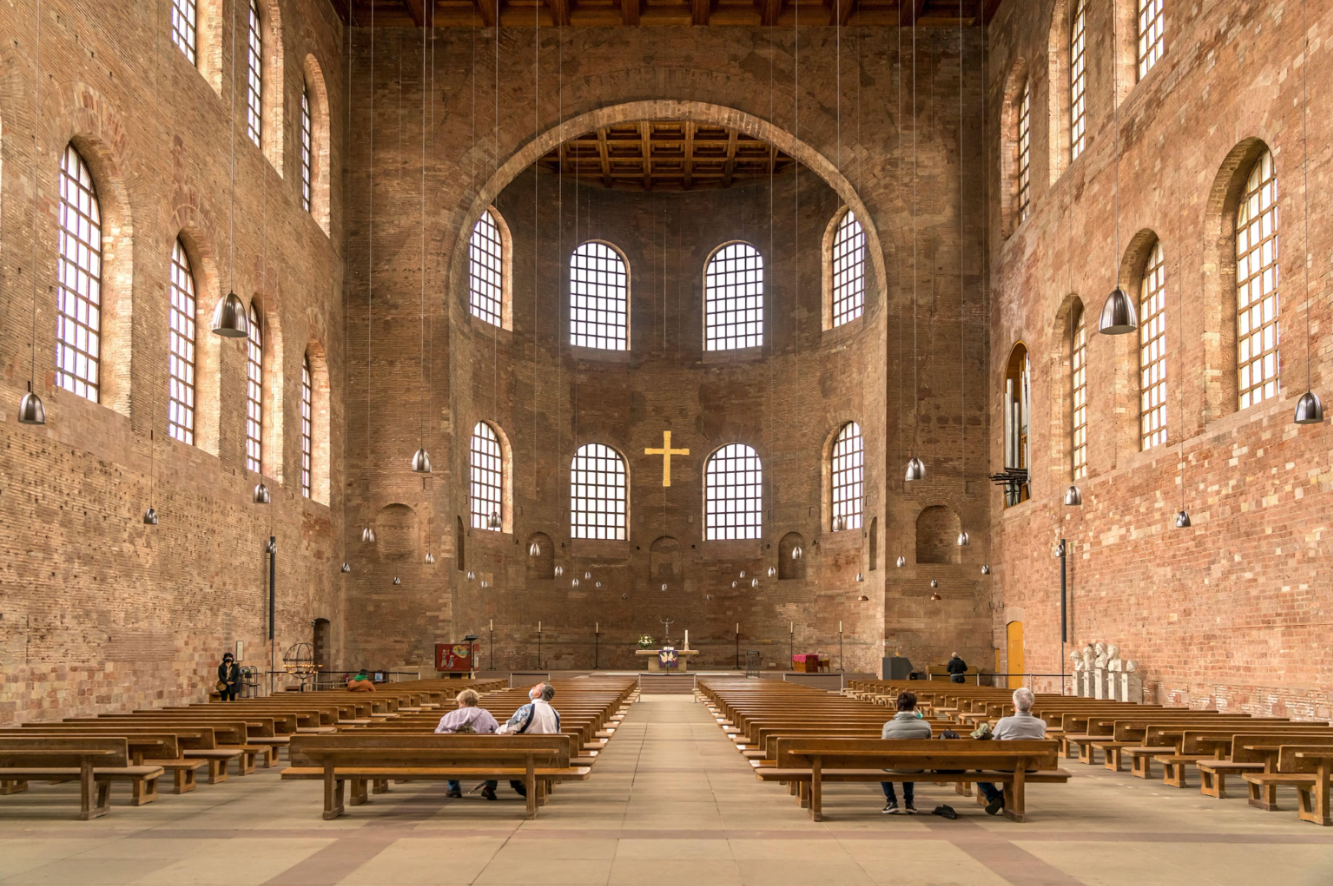 View of the inside of a large cathedral from the entrance. There are pews on the left and right with several people sitting in them. There is an altar in the back and a gold cross hanging halfway from the ceiling.