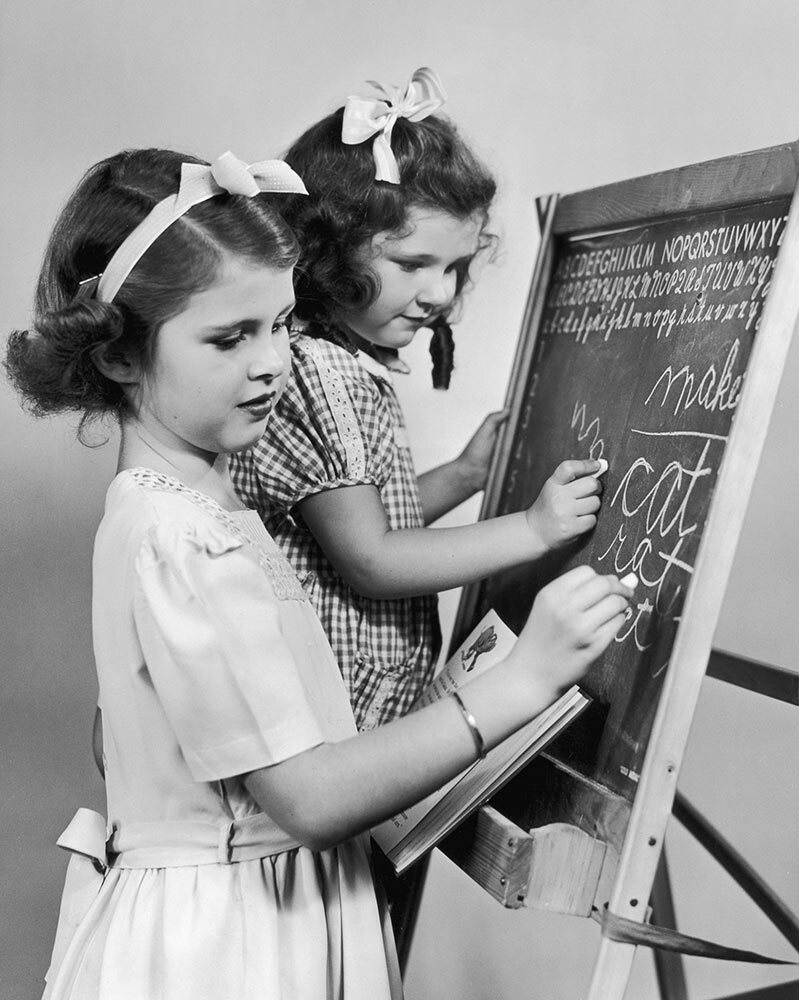 two young girls practice cursive on a chalkboard in 1953