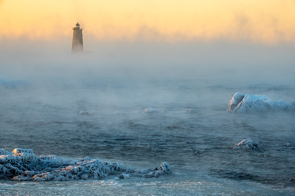 Heavy fog is seen over the ocean, it covers a portion of a lighthouse on the horizon.