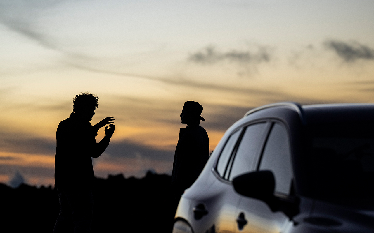 A photo of a silhouette of two men talking next to a car, the sun setting in the background.