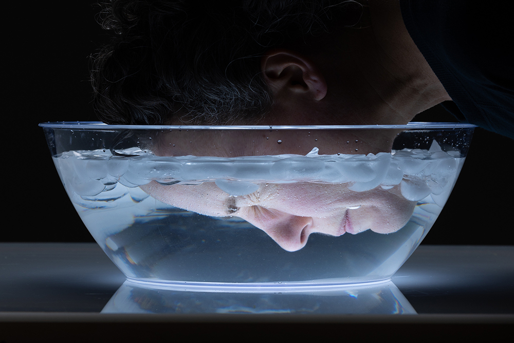 A woman's face, seen from the side, as she submerges her face in a bowl of ice water