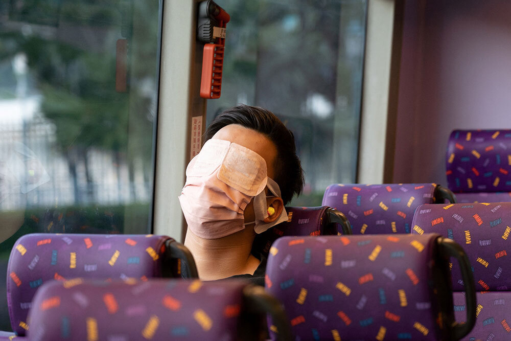 A man sits on a bus wearing an eye mask, ear plugs, and a mask for his mouth.