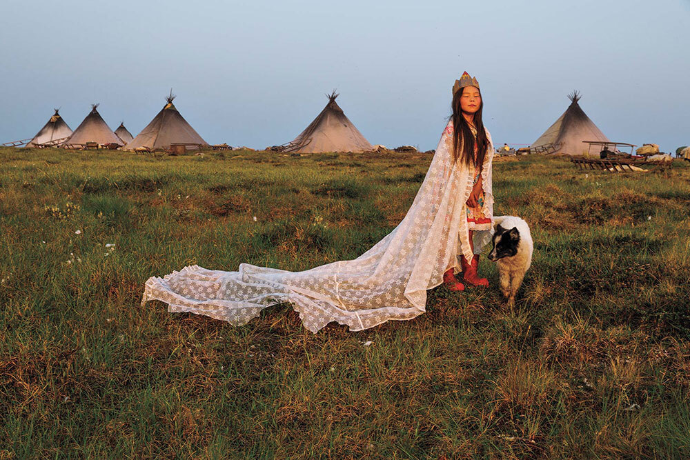 A girl wears a long cape and crown in front of a row of teepees