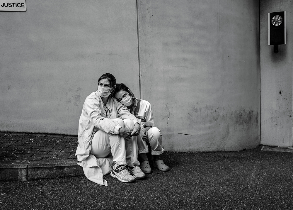 Two nurses, wearing scrubs and face masks sitting together at the end of a sidewalk. Both nurses appear exhausted as one rests her head on the other's shoulder