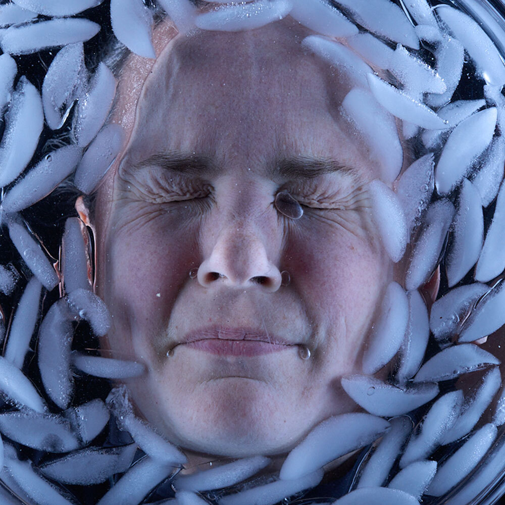 A woman's face, seen from below, surrounded by ice cubes as she plunges her face into a bowl of ice water