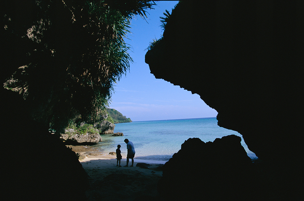 A father and son stand in silhouette along Okinawa Island beach, framed by eroded rocks and tropical foliage.