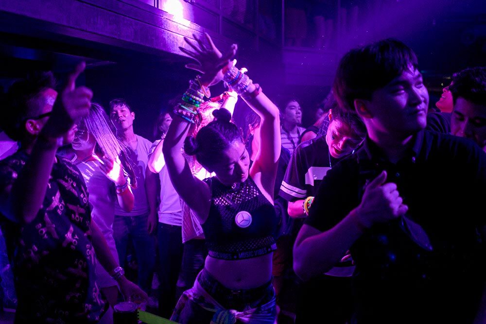 A woman dancing, with her arms above her head, under moody, purple lights, amongst a crowd.