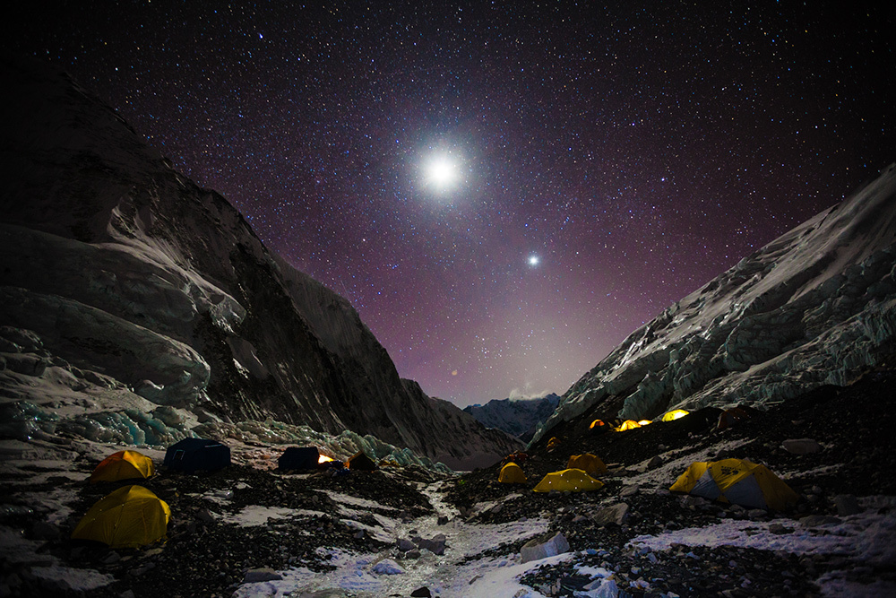 A star-filled sky and bright moon shine over dozens of tents set up at a rocky and icy campsite on Everest.