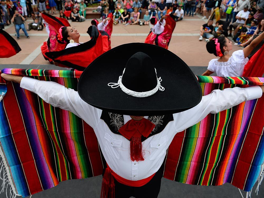 A member of a dance troop stands with arms outstretched, supporting a colorful sarape, with dancers behind him.