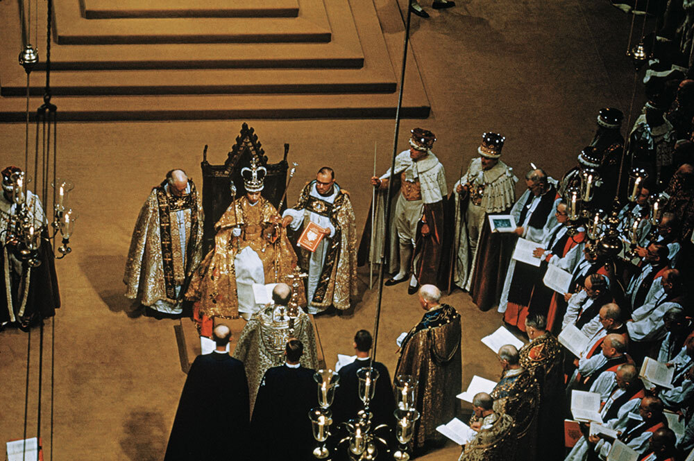 The queen wears a crown and gown surrounded by men in robes