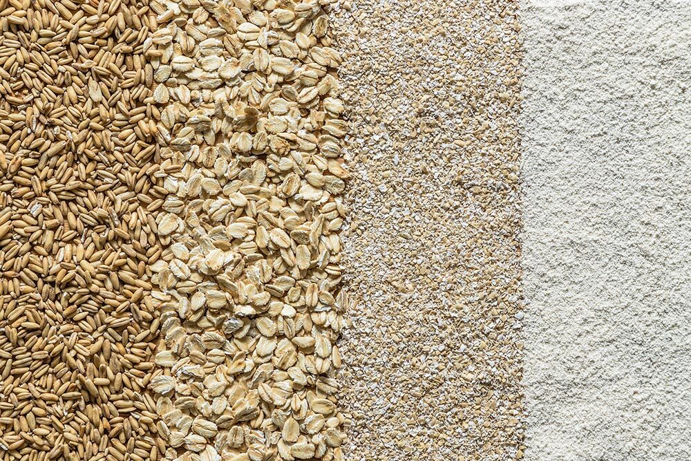 Top view of various types of oats arranged in vertical strips, showing whole oat grains, oat flakes, oat bran and oat meal/flour.