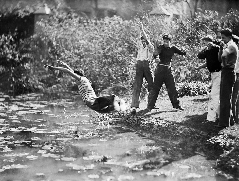 a laughing crowd of teenage boys throws another boy into a pond