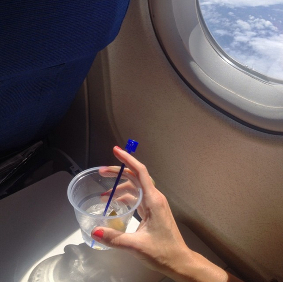 A manicured hand holding a cocktail in a plastic cup while sitting beside an airplane window.