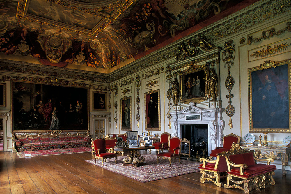 A large, elaborate room with wood floors, high and painted gold adorned ceilings, velvet and gold furniture, paintings on the wall, and a large fireplace.