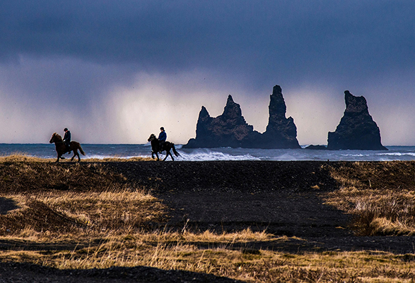 Two horseback riders travel down the southern coast of Iceland, passing the Reynisdrangar sea stacks.