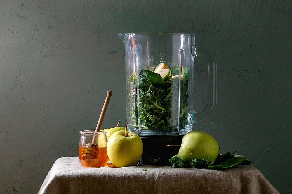 Ingredients for green spinach kale apple honey smoothie in glass blender on linen table cloth with ingredients above.