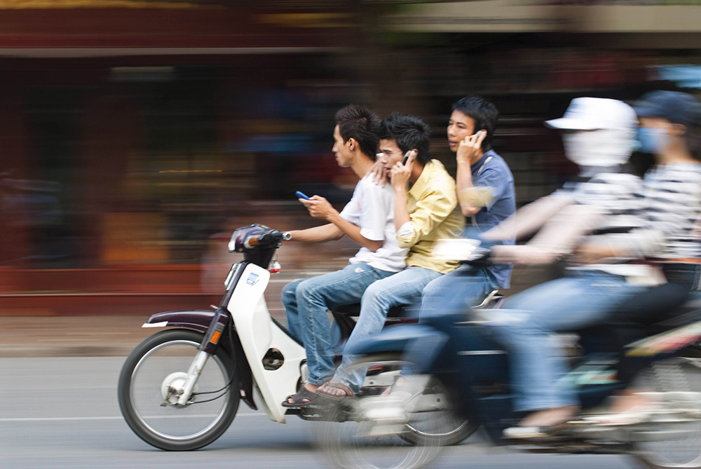 Three young men using their phones while riding on a scooter.