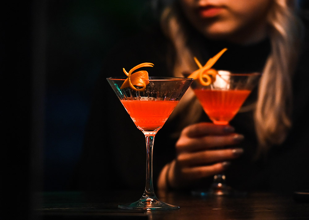 A woman drinking pink cocktail from a martini glass, garnished with a twist of orange peel.