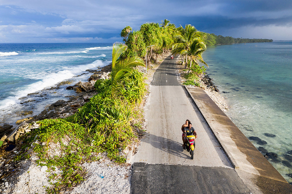 Two people ride on a motorbike down a narrow road surrounded by blue water; on the left is the Pacific Ocean and the right, a lagoon