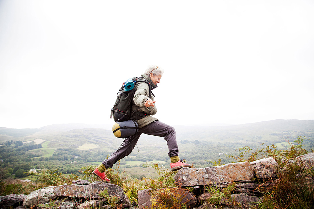 Older woman, with a backpacking back pack, a light grey coat and dark grey pants, happily trekking the mount